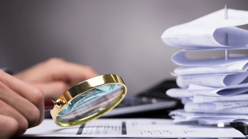 Close-up of a hand holding a gold-rimmed magnifying glass over documents, with a stack of papers and a calculator in the background.