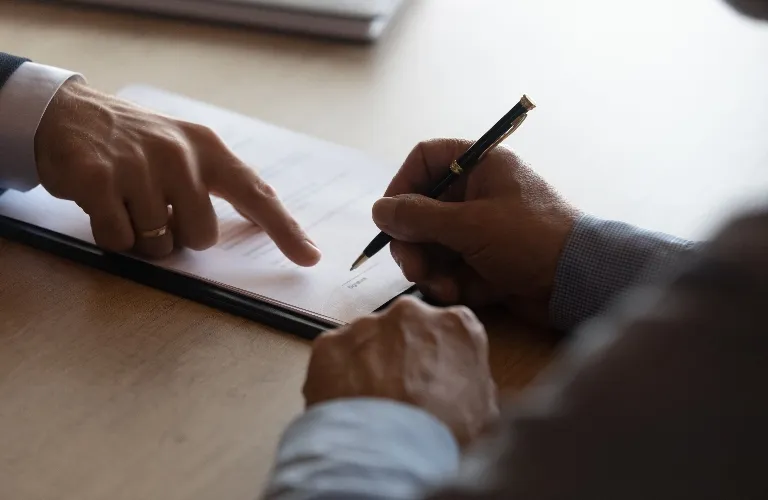 Two individuals engaged in signing a document on a wooden table, one pointing while the other holds a pen.