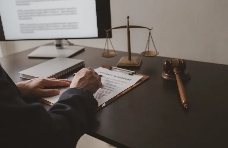 Person signing a contract agreement on a clipboard at a desk with legal scales and gavel.