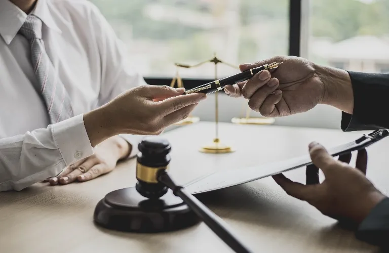 Two professionals exchanging a black and gold pen over a desk with legal gavel and scales in the background.