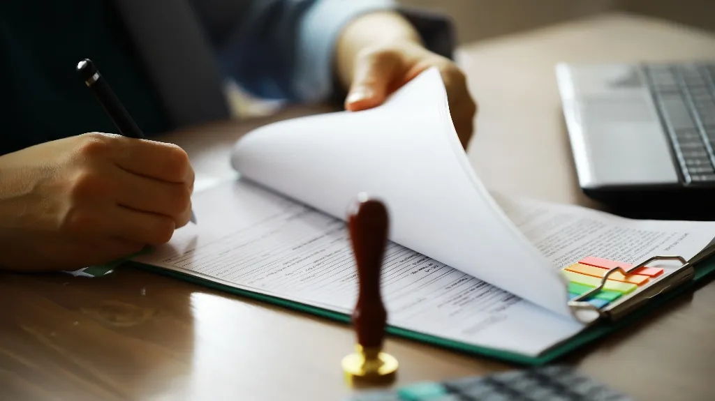 Person signing a document on a clipboard with colorful tabs near a laptop and stamp.