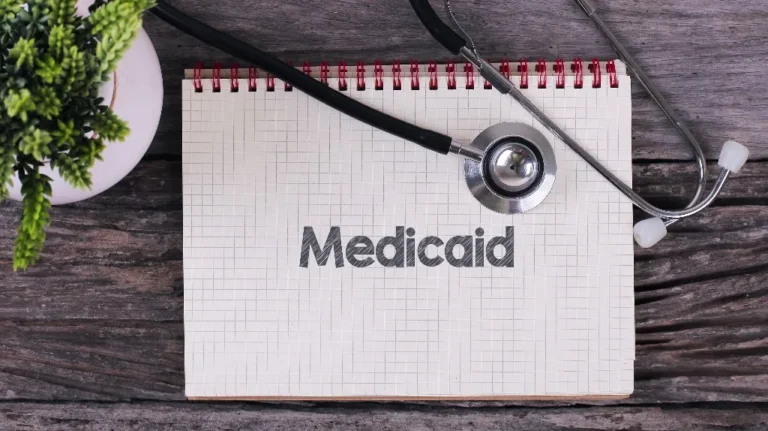 Spiral notebook with grid paper and the word "Medicaid" printed, black stethoscope, and small green plant on wooden surface.