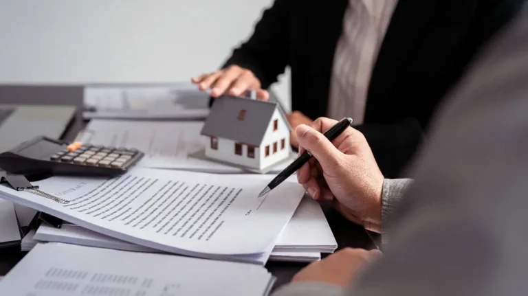 Two professionals reviewing and signing real estate documents with a miniature house model and calculator on the table.