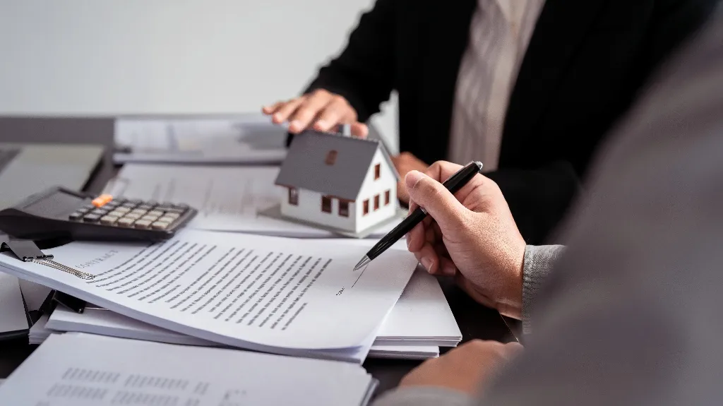 Two professionals reviewing and signing real estate documents with a miniature house model and calculator on the table.