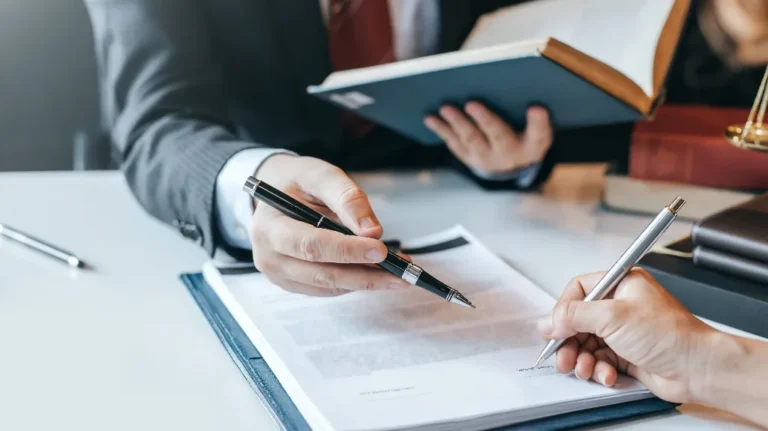 Two professionals in business attire reviewing and signing a document at a white desk.