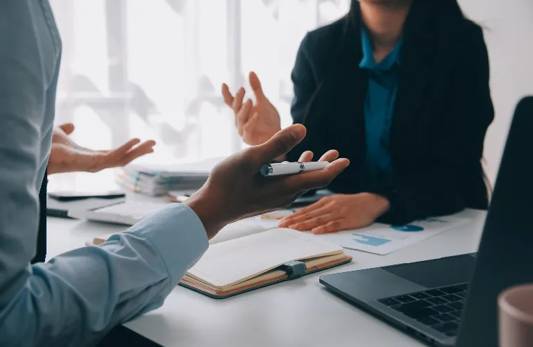 Two professionals engaged in discussion at a desk with documents, notebook, and laptop.