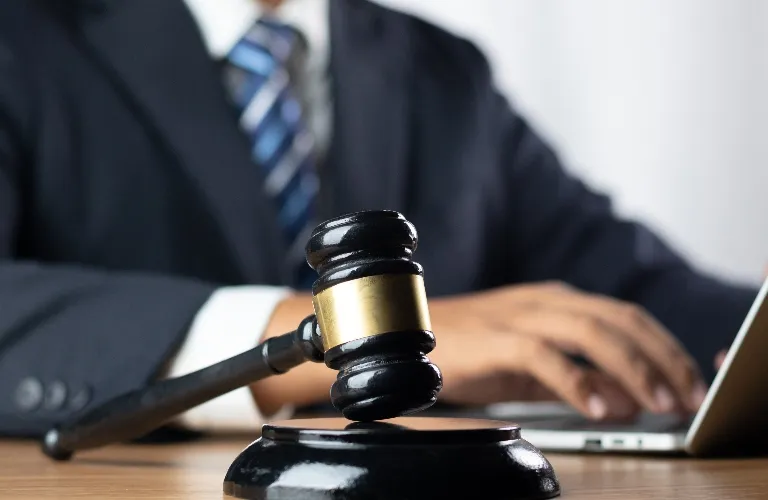 Close-up of a black judge's gavel with gold band on wooden desk, businessman typing on laptop in background.