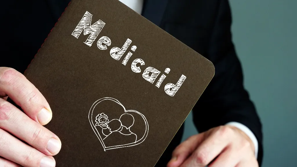 Person in dark suit holding a brown booklet labeled "Medicaid" with a heart and family illustration.