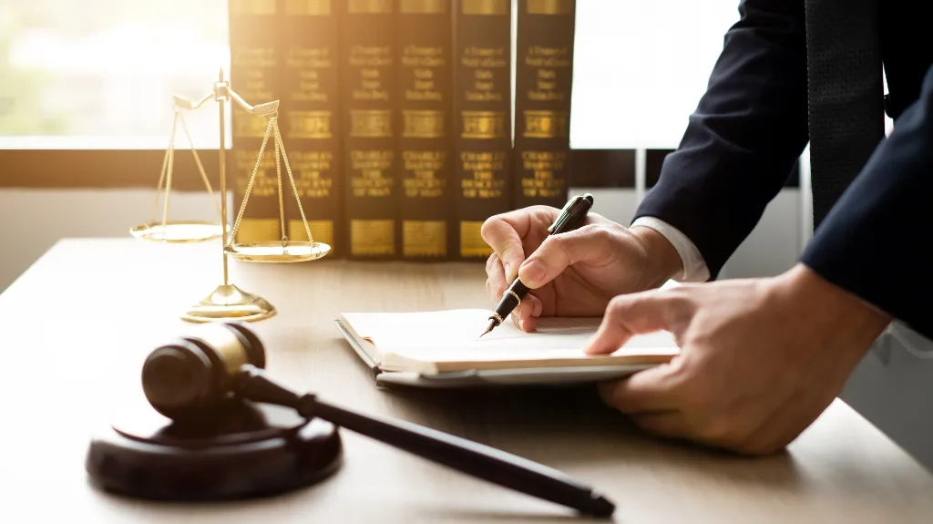 Person in dark suit writing on a notepad at a desk with legal books, scales of justice, and a gavel.