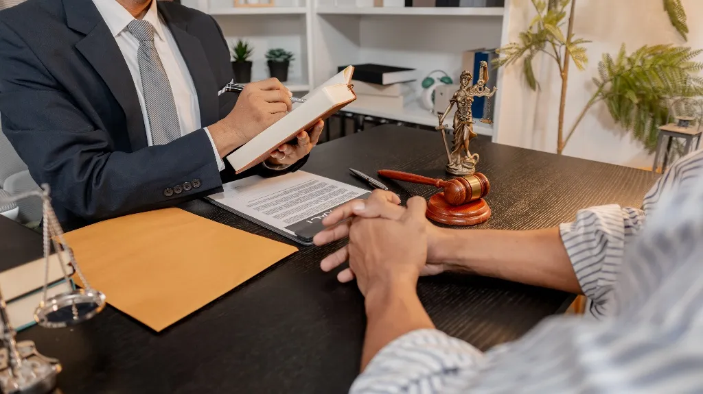 Professional consultation between a lawyer in suit and a client at a desk with legal documents, gavel, and Lady Justice statue.