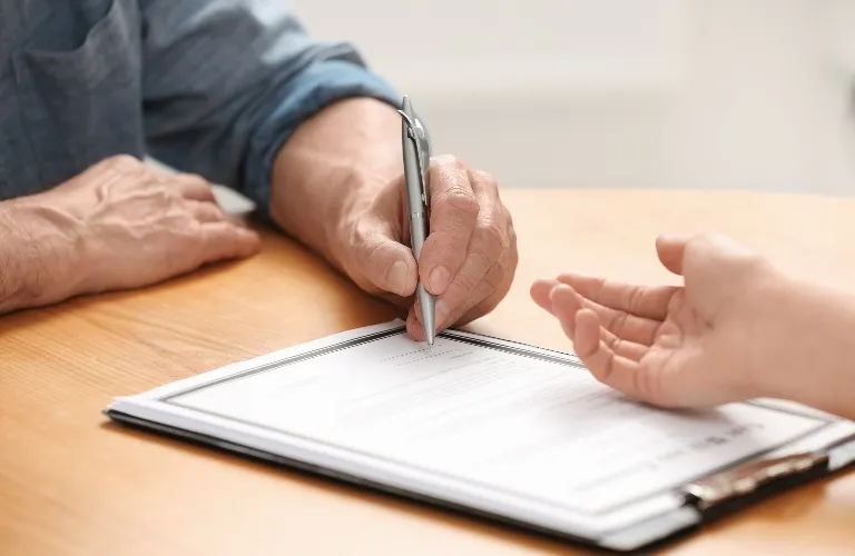 Close-up of two people at a wooden table, one signing a document with a pen.