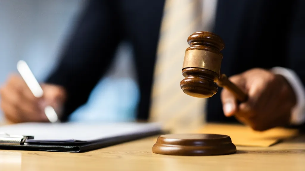 Close-up of a wooden judge's gavel held by a person in a suit, with a clipboard and pen in the background.
