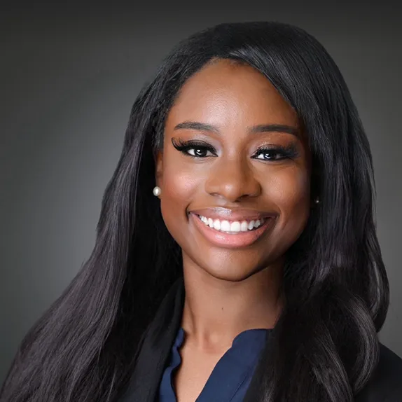 Professional portrait of a smiling woman with long black hair and pearl earrings against a gray background.