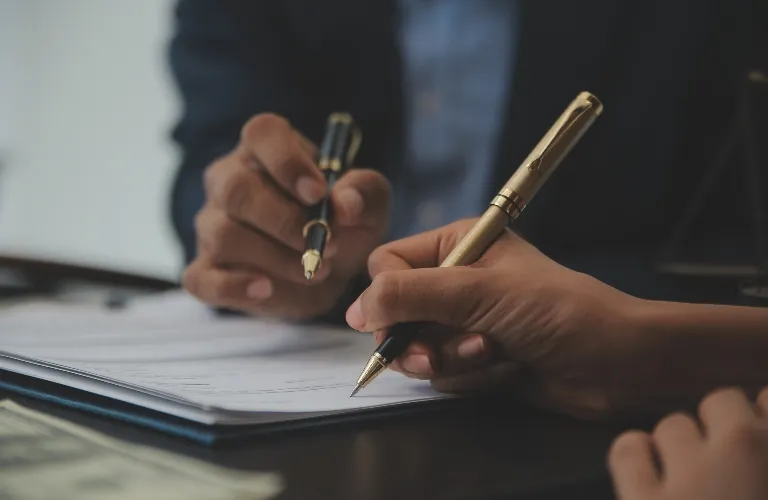 Two individuals holding pens over a document on a dark table, engaged in signing or reviewing.