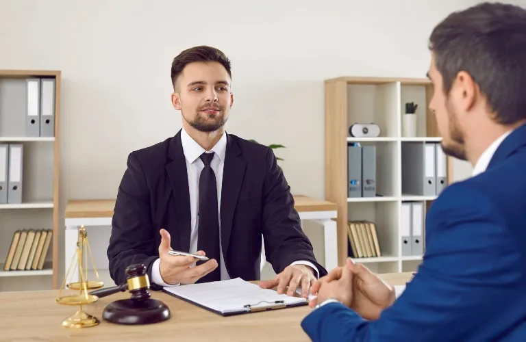 Two professionally dressed men engaged in discussion at an office desk with legal scales and a gavel.