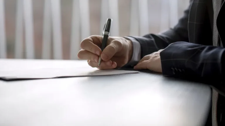 Close-up of a person in a dark suit signing a document with a black pen on a white table.