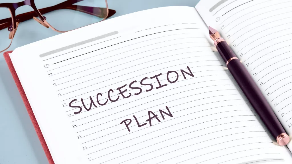 Open notebook with "Succession Plan" written in bold letters, black fountain pen, and eyeglasses.